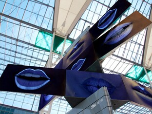 Der Meta Twist Tower, der aus eckigen Komponenten aus Metall besteht. Auf der schwarzen Grundfarbe des Objekts sind blaue, geöffnete Münder in fluoreszierender Farbe abgebildet.