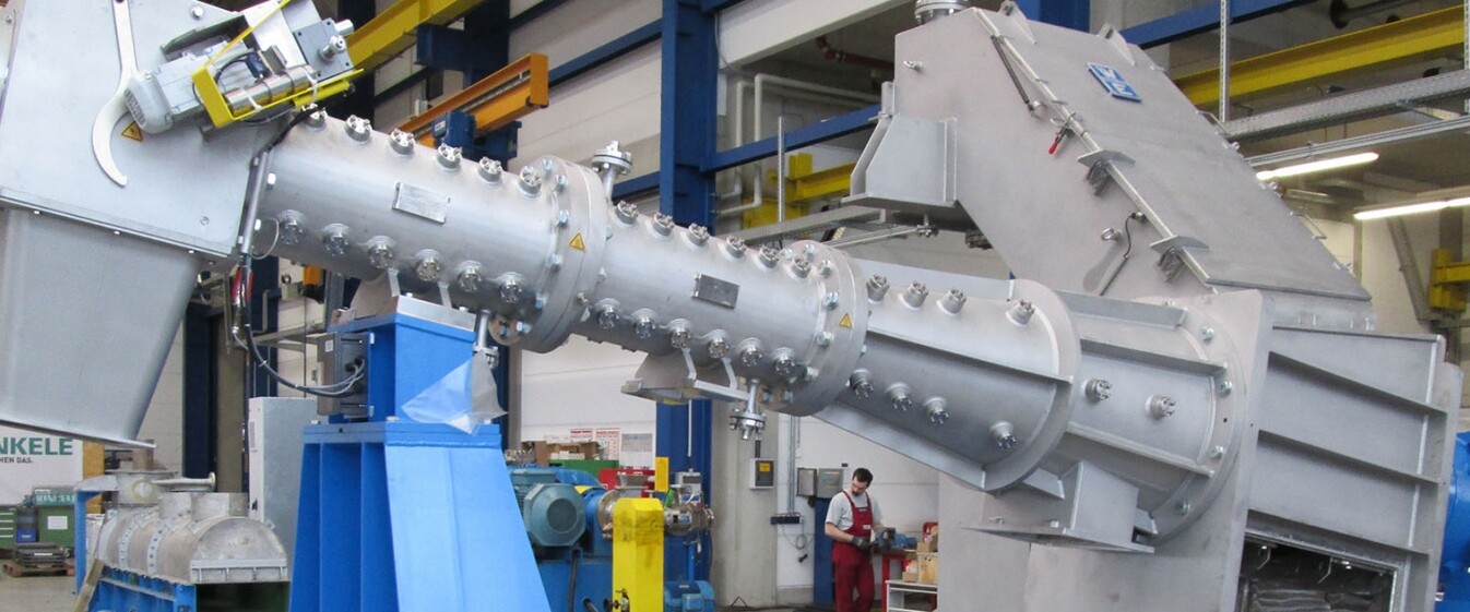 Große silberne Maschine, die ein Teil eines Extruders ist. Im Hintergrund arbeiten Personen in einer großen Halle.
