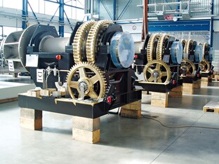 Uhrwerke mit großen Zahnrädern und mechanischen Komponenten, auf Holzböcken in einer Produktionshalle platziert. Mehrere identische Maschinen in einer Reihe, in einer hellen Fabrikumgebung.