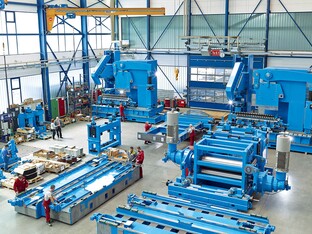 Die Montage einer blauen Spiralrohrmaschine. Die einzelnen Teile liegen nebeneinander in einer großen Halle, in der Personen arbeiten.