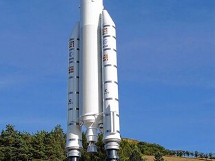 Modell der Rakete Ariane 5 in einer Außenumgebung, stehend auf einem grünen Gelände bei blauem Himmel. Das Modell zeigt die charakteristischen zwei seitlichen Booster und den zentralen Raketenkörper.