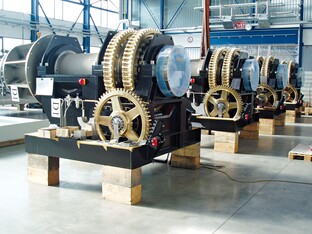 Uhrwerke mit großen Zahnrädern und mechanischen Komponenten, auf Holzböcken in einer Produktionshalle platziert. Mehrere identische Maschinen in einer Reihe, in einer hellen Fabrikumgebung.