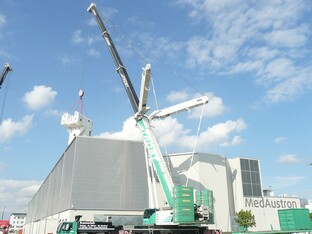 Ein Kran hebt ein großes Konstrukt in nach oben, während ein weiteres, industrielles Gebäude im Hintergrund sichtbar ist.