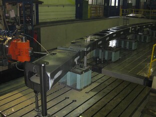 Industrielle Nietvorrichtung mit langer Metallstruktur auf Stützen in einer Werkshalle, mit einem orangefarbenen Bearbeitungswerkzeug im Vordergrund.