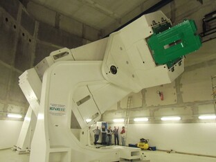 Eine große, weiße Maschine, die in einem sehr hohen Raum steht. Im Hintergrund sind Personen, welche deutlich kleiner, als die Maschine sind.