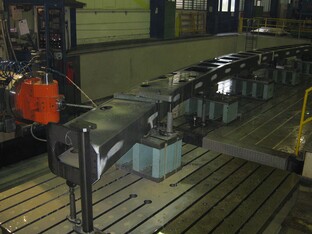 Industrielle Nietvorrichtung mit langer Metallstruktur auf Stützen in einer Werkshalle, mit einem orangefarbenen Bearbeitungswerkzeug im Vordergrund.