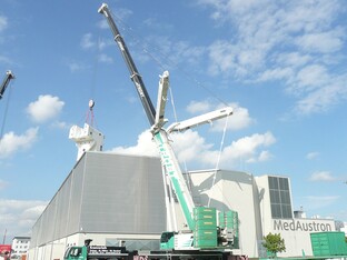 Ein Kran hebt ein großes Konstrukt in nach oben, während ein weiteres, industrielles Gebäude im Hintergrund sichtbar ist.
