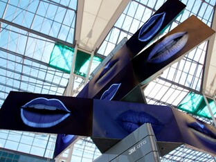 Der Meta Twist Tower, der aus eckigen Komponenten aus Metall besteht. Auf der schwarzen Grundfarbe des Objekts sind blaue, geöffnete Münder in fluoreszierender Farbe abgebildet.