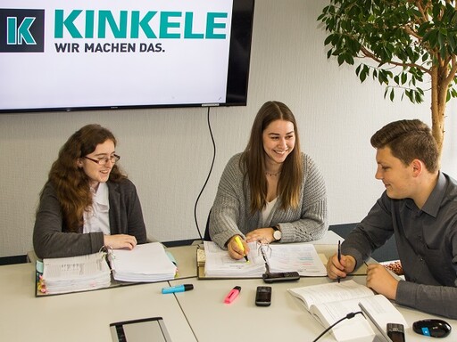 Zwei Frauen und ein Mann, alle lächelnd, sitzen an einem Konferenztisch mit Ordnern. Im Hintergrund, auf einem Bildschirm, das Logo von Kinkele.