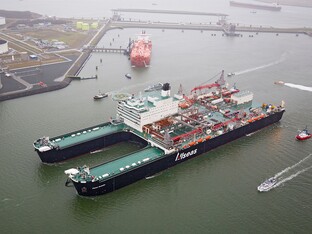 Großes Frachtschiff, welches beladen ist. Im Hintergrund ist eine Art Hafen zu sehen mit Anlegestellen.