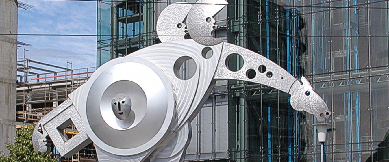 Eine silberne Statue mit einem Pferdekopf und zwei Halbmonden auf dem Rücken. Im Hintergrund eine moderne urbane Umgebung.